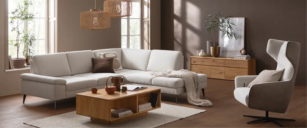 Beiges Ecksofa vor hohen Fenstern, davor niedriger Couchtisch aus Holz auf hellem Teppich, umgeben von Pflanzen und Sideboard im Wohnzimmerstil.
